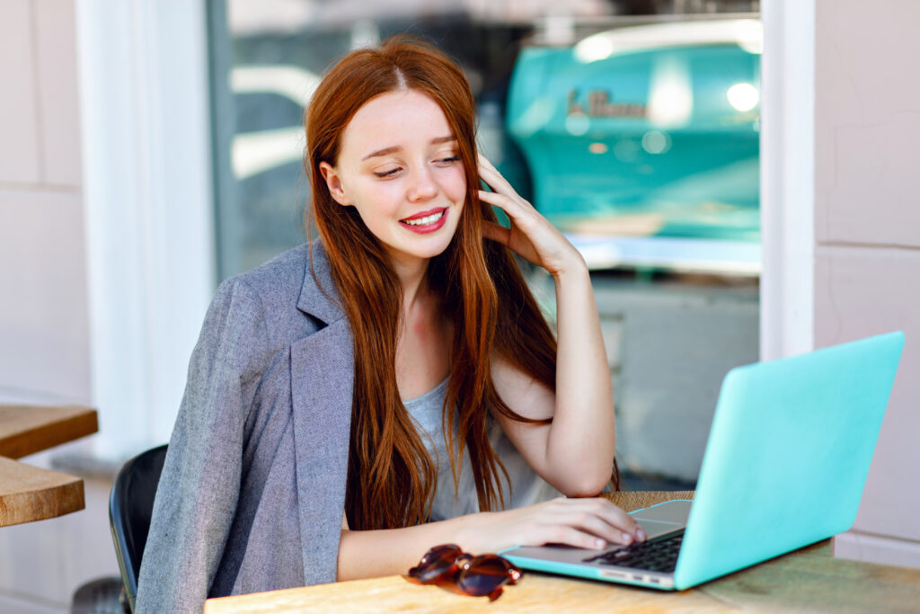 A young businesswoman working on her laptop at an outdoor café, smiling while engaging in guest posting for local blogs. She is dressed in a casual yet professional outfit, representing modern digital marketing and networking strategies for business growth.